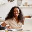 Attractive stylish young dark skinned female in beige shirt sitting at kitchen table, using laptop, calculating budget, planning vacations, smiling happily. Self employed black woman working from home